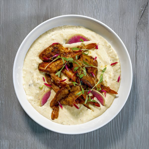 Shrimp and grits with microgreens and radish slices in a white bowl on a wooden table.