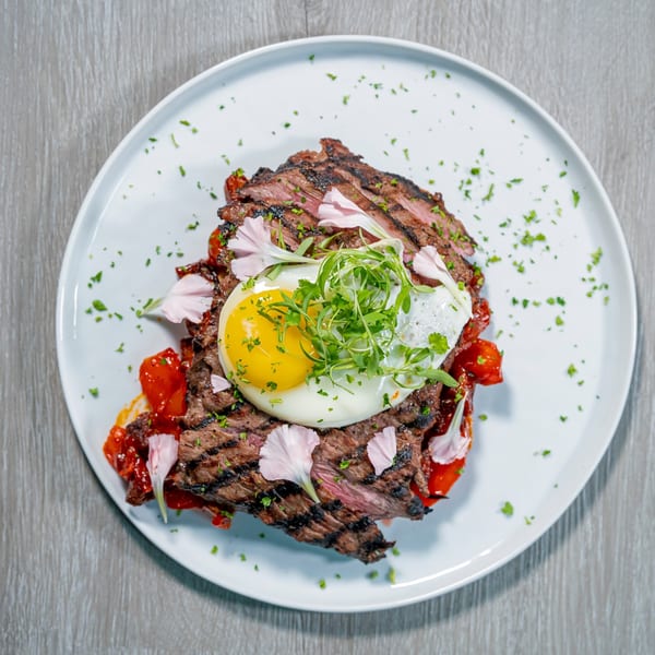 Grilled steak with a fried egg, red peppers, and microgreens, garnished with edible flowers.