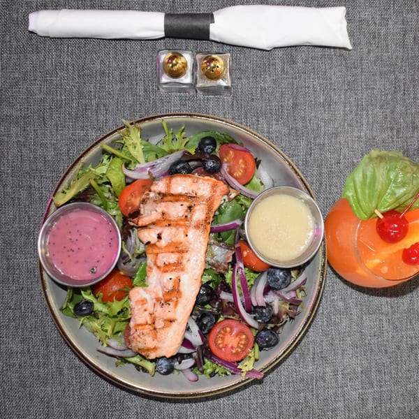 Grilled salmon salad with mixed greens, tomatoes, blueberries, red onion, and two dressings, with a cocktail.